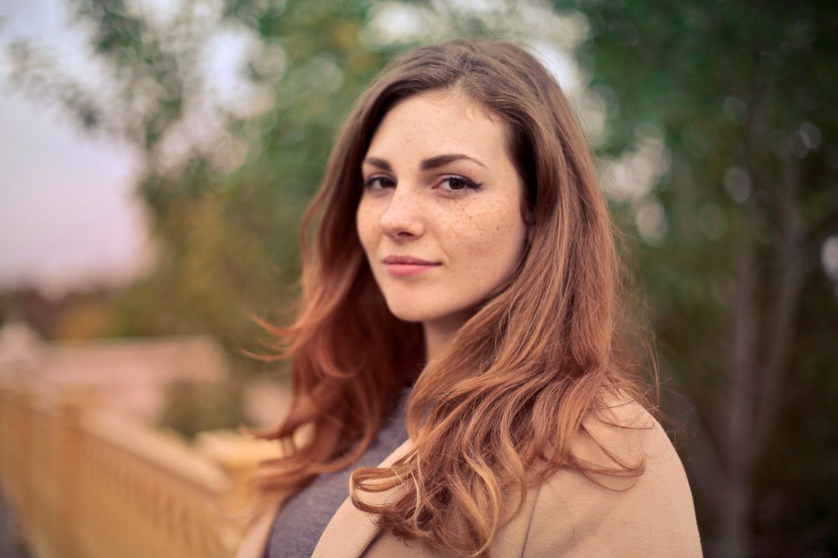 A woman in a tan coat looking towards the camera outdoors